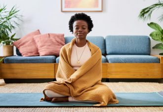 An attractive young Black woman sitting on a yoga mat in her lounge at home meditating.